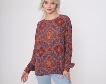 May include: A long-sleeved, loose-fitting blouse with a colorful paisley print in shades of orange, red, blue, and purple. The blouse has a round neckline and a relaxed fit.