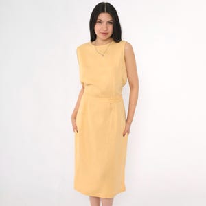 Peut inclure: Une robe sans manches, mi-longue, de couleur jaune pâle. La robe a une silhouette simple et ajustée et un décolleté arrondi.