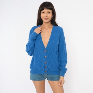 May include: A bright blue V-neck cardigan sweater featuring a cable knit pattern throughout the body and sleeves. The garment has a button-down front closure with five matching blue buttons and ribbed cuffs and a ribbed hem at the bottom edge.