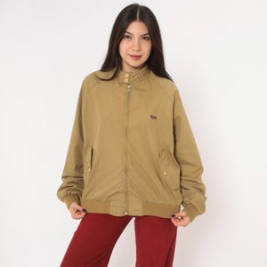 May include: A tan zip-up jacket featuring a high collar with two buttons, long sleeves with ribbed cuffs, and two front pockets with button closures. The jacket has a small rectangular logo patch on the left chest and a ribbed hem at the waist.