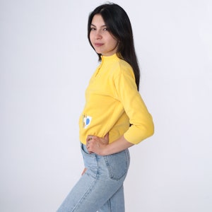 May include: A yellow long-sleeved sweater with a blue and yellow graphic design. The sweater has a mock neck and a zip at the neck.