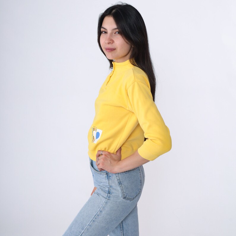 May include: A yellow long-sleeved sweater with a blue and yellow graphic design. The sweater has a mock neck and a zip at the neck.
