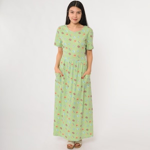 May include: A light green maxi dress with a round neckline and short sleeves. The dress features a pattern of small, colorful illustrations. It has two front pockets and a flowing skirt, creating a casual and comfortable look.