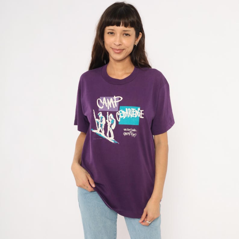 May include: A purple t-shirt with the words "CAMP CEDARIDGE" in white and teal. The graphic shows two figures on a surfboard. The shirt is a crew neck and has short sleeves.