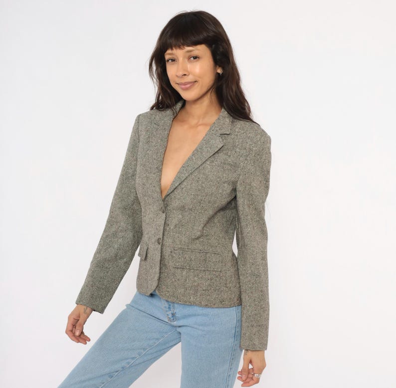 May include: A gray tweed blazer with a single-button closure and two front pockets. The blazer has a notched lapel and long sleeves. The model is wearing light blue jeans. The blazer is a classic style.