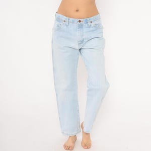 May include: Light blue denim jeans with a classic five-pocket design. The jeans have a straight leg cut and a button and zipper closure. The light wash denim provides a vintage look. The jeans are worn by a person.