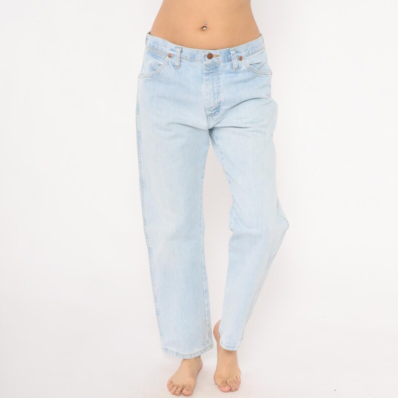 May include: Light blue denim jeans with a classic five-pocket design. The jeans have a straight leg cut and a button and zipper closure. The light wash denim provides a vintage look. The jeans are worn by a person.