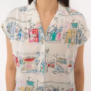 May include: A short-sleeved button-down shirt in off-white featuring a colorful hand-drawn style print of city buildings, houses, and street scenes in shades of red, blue, yellow, and pink with black ink outlines and a classic collar.