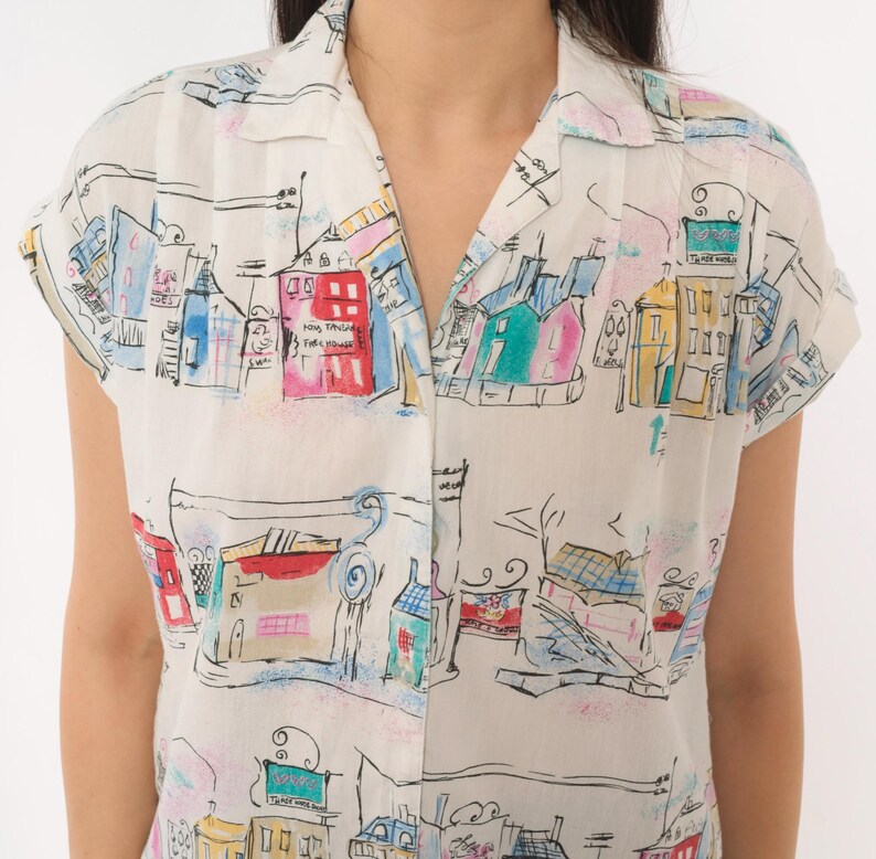 May include: A short-sleeved button-down shirt in off-white featuring a colorful hand-drawn style print of city buildings, houses, and street scenes in shades of red, blue, yellow, and pink with black ink outlines and a classic collar.