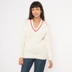 May include: Cream-colored cable knit sweater with a V-neckline trimmed in pink. The long-sleeved sweater is made of a thick knit material. The model is wearing blue jeans.
