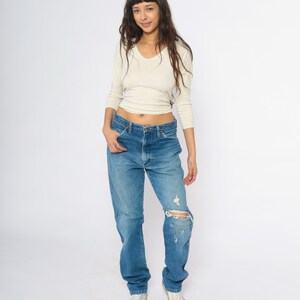 May include: A woman in a cream V-neck top and blue jeans. The jeans are distressed with a ripped knee. She is wearing white trainers. The outfit is casual and relaxed.