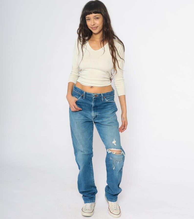 May include: A woman in a cream V-neck top and blue jeans. The jeans are distressed with a ripped knee. She is wearing white trainers. The outfit is casual and relaxed.