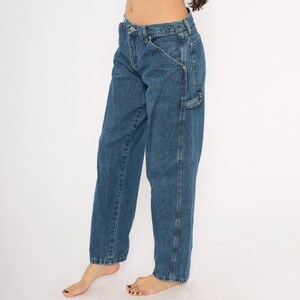 May include: Blue denim carpenter pants with a relaxed fit. The jeans feature a classic five-pocket design, a hammer loop, and a utility pocket. The pants have a straight leg and a medium wash.