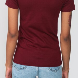 May include: A maroon t-shirt with short sleeves and a crew neck. The t-shirt is worn by a person wearing light blue jeans.