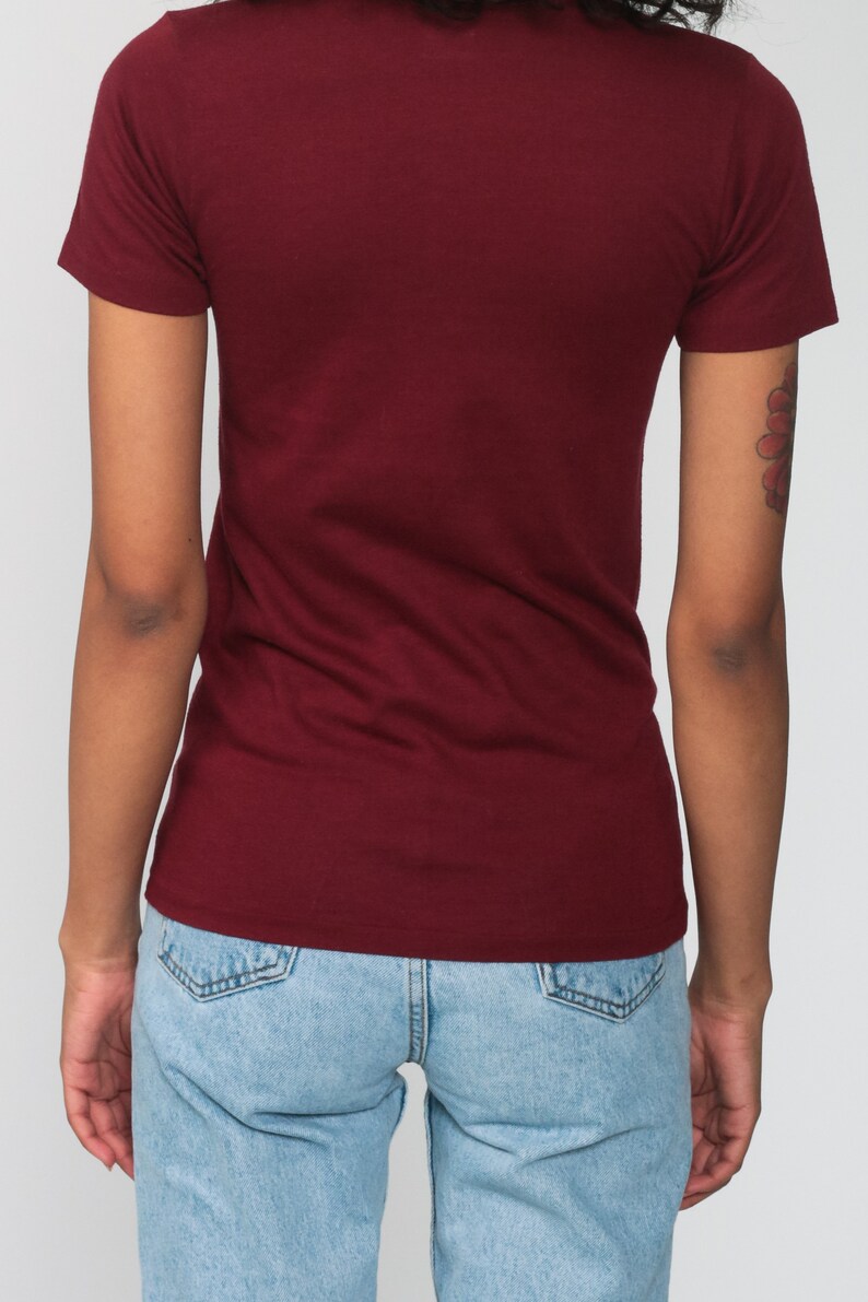May include: A maroon t-shirt with short sleeves and a crew neck. The t-shirt is worn by a person wearing light blue jeans.