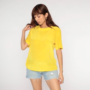 以下が含まれることがあります： 鮮やかな黄色の半袖Tシャツ、丸首。柔らかく流れるような生地です。モデルはライトブルーのデニムショートパンツを着用しています。背景は無地の白です。
