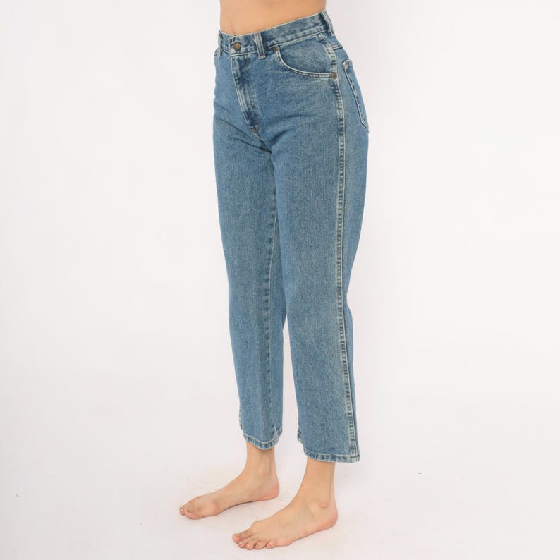 May include: Light blue denim jeans with a high waist and straight leg. The jeans have a classic five-pocket design and a button and zipper closure. The jeans are cropped at the ankle.