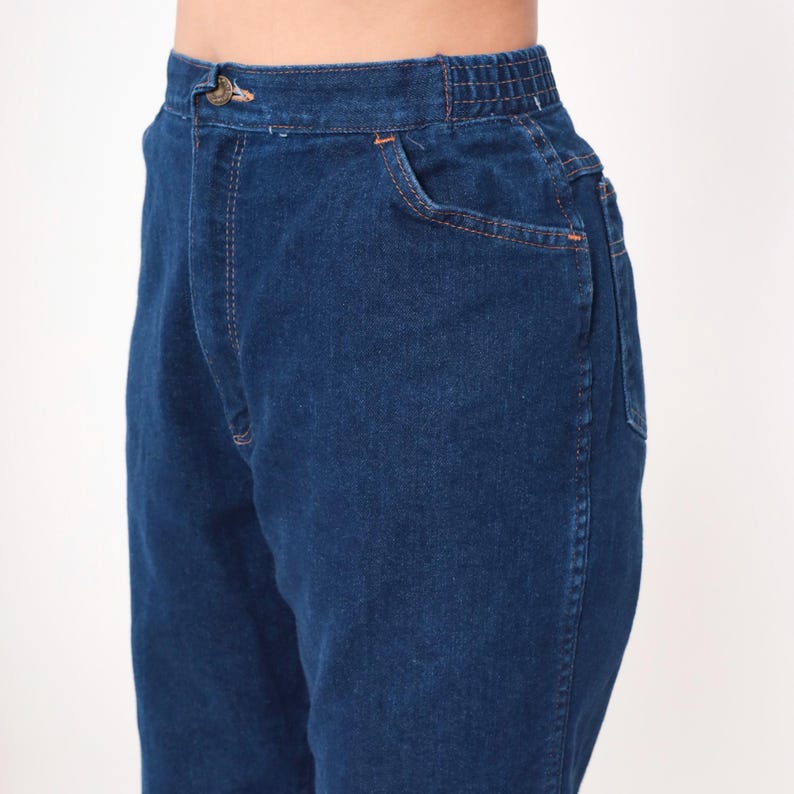 K&ouml;nnte beinhalten: Nahaufnahme einer dunkelblauen Jeanshose. Die Hose hat einen Knopf und einen elastischen Bund. Die Vordertasche und die N&auml;hte sind mit orangefarbenen N&auml;hten versehen. Die Hose ist aus robustem Denim-Material.