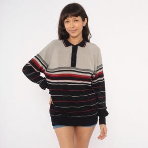 May include: A long-sleeved knit polo shirt featuring a colour-blocked design with a light grey upper section and a black lower section. The garment includes horizontal stripes in red, white, and black across the chest and sleeves, with a three-button placket.