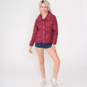 May include: A burgundy quilted jacket with a high collar and silver buttons. The jacket is paired with denim shorts and white sneakers. The outfit is set against a white background, showcasing a casual, everyday style.