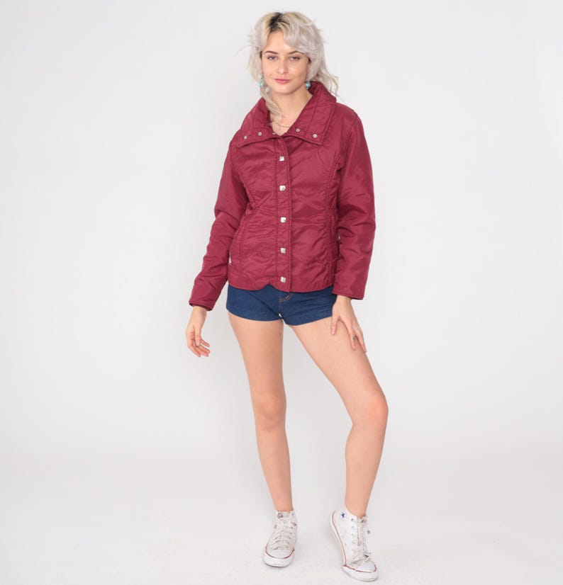 May include: A burgundy quilted jacket with a high collar and silver buttons. The jacket is paired with denim shorts and white sneakers. The outfit is set against a white background, showcasing a casual, everyday style.