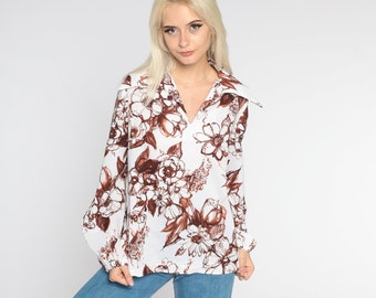 Puede incluir: Una blusa blanca de manga larga con estampado floral marrón. La blusa tiene cuello y escote en V.