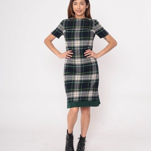 May include: A short-sleeved plaid dress in shades of navy, green, and cream. The dress features a knee-length hemline with a green border and is paired with black boots. The overall style is classic and preppy.