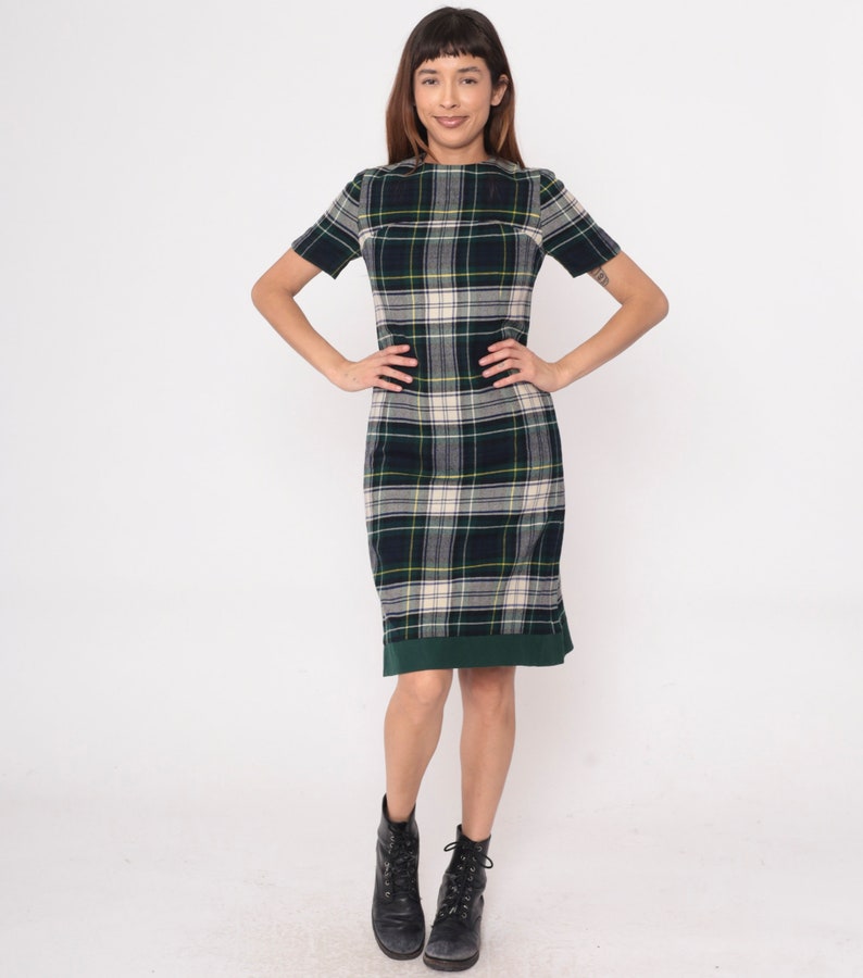 May include: A short-sleeved plaid dress in shades of navy, green, and cream. The dress features a knee-length hemline with a green border and is paired with black boots. The overall style is classic and preppy.