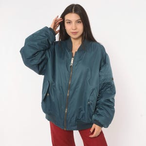 May include: A teal bomber jacket featuring a front metal zipper closure, ribbed collar, cuffs, and hem. The jacket includes two side pockets with snap button closures and a utility pocket on the left sleeve, constructed from a smooth, solid fabric.