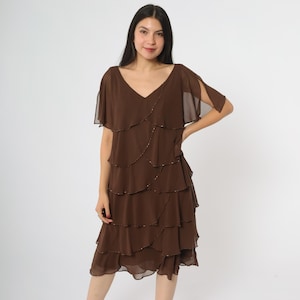 Peut inclure: Robe marron à plusieurs niveaux avec un décolleté en V et des manches courtes à volants. La robe présente un design fluide et superposé avec de subtils accents perlés. La robe, de longueur genou, est faite d'un tissu léger et semi-transparent.