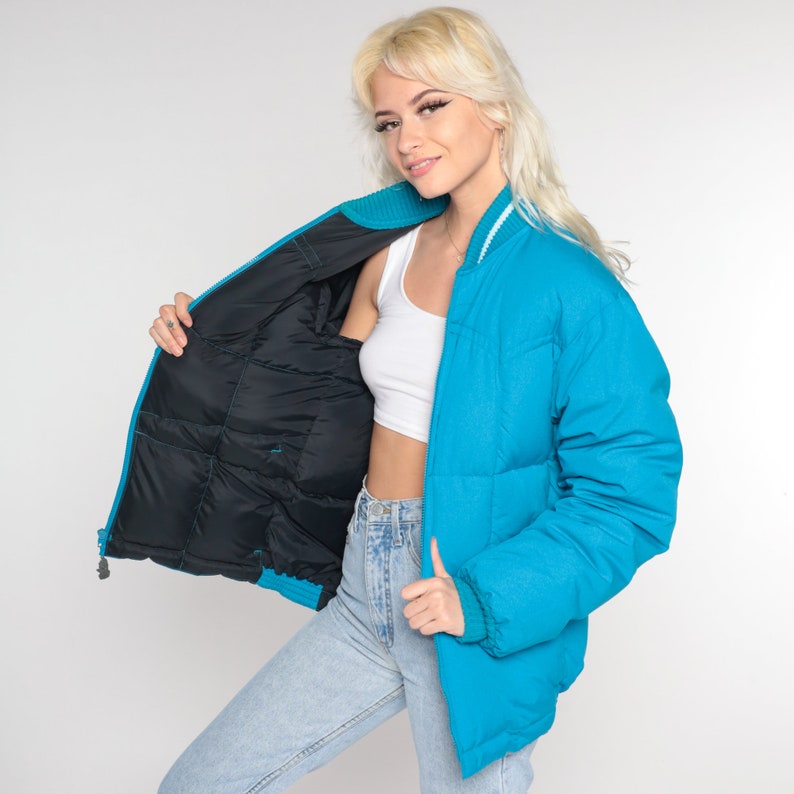 May include: A turquoise blue puffer jacket with a black quilted lining. The jacket has a zipper closure and a white trim around the collar.