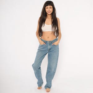 Peut inclure: Une femme porte un haut court blanc et un jean en denim bleu clair. Le jean a une conception classique &agrave; cinq poches et une coupe d&eacute;contract&eacute;e. Le mod&egrave;le se tient devant un fond blanc.