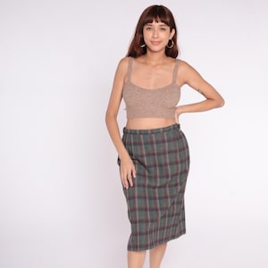 May include: A beige knit crop top with wide straps and a green, red, and gray plaid skirt. The skirt is knee-length and has a high waist. The outfit is shown against a white background.