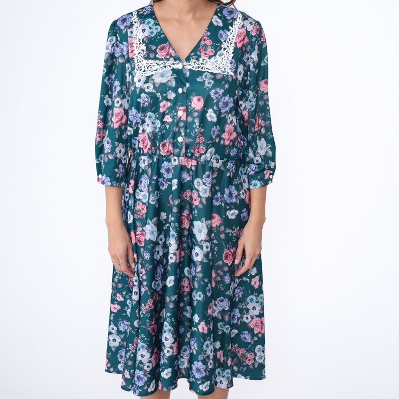 K&ouml;nnte beinhalten: Ein blaues Midi-Kleid in Teal mit einem Blumendruck mit rosa, wei&szlig;en und blauen Blumen. Das Kleid hat einen wei&szlig;en Spitzenbesatz am Ausschnitt und eine Knopfleiste vorne.