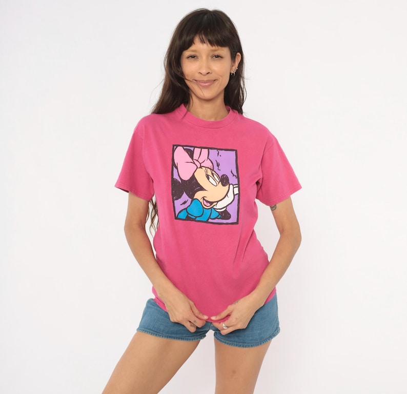 May include: A short-sleeved, crew-neck t-shirt in a vibrant magenta pink colour. The front features a square graphic print of Minnie Mouse wearing a pink bow and a blue dress, framed by a thick black border against a light purple background.
