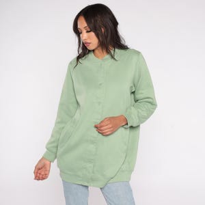 May include: A light green button-up jacket with long sleeves. The jacket has a round collar and two front pockets. The fabric appears to be a soft, comfortable material. The jacket is worn over blue jeans.