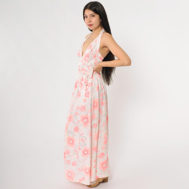 May include: A halter-neck maxi dress with a floral pattern in shades of pink and white. The dress features a deep V-neckline and a gathered waist detail. The long skirt flows to the floor.