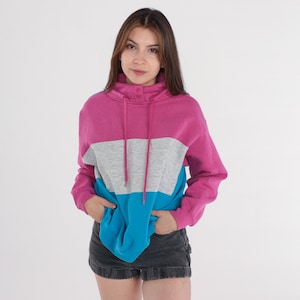 May include: A color-block sweatshirt in pink, gray, and blue. The sweatshirt has a high collar with two buttons and a drawstring. The sleeves are long, and the person is wearing black denim shorts. The background is white.