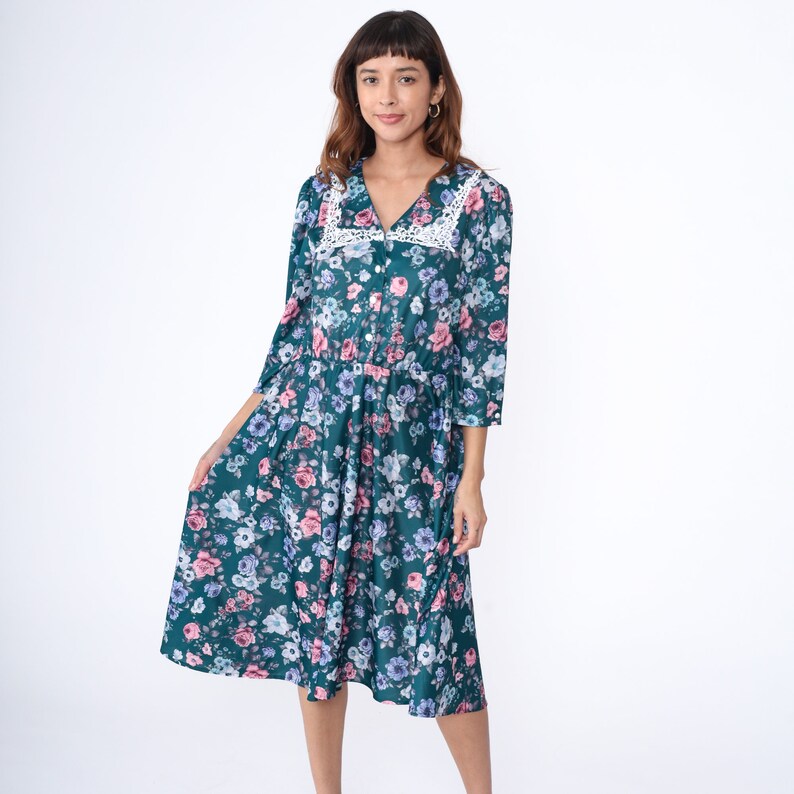 K&ouml;nnte beinhalten: Ein blaues Midi-Kleid mit einem wei&szlig;en Spitzenkragen und einem Blumenmuster. Das Kleid hat einen Knopfleisten vorne und 3/4-&Auml;rmel.
