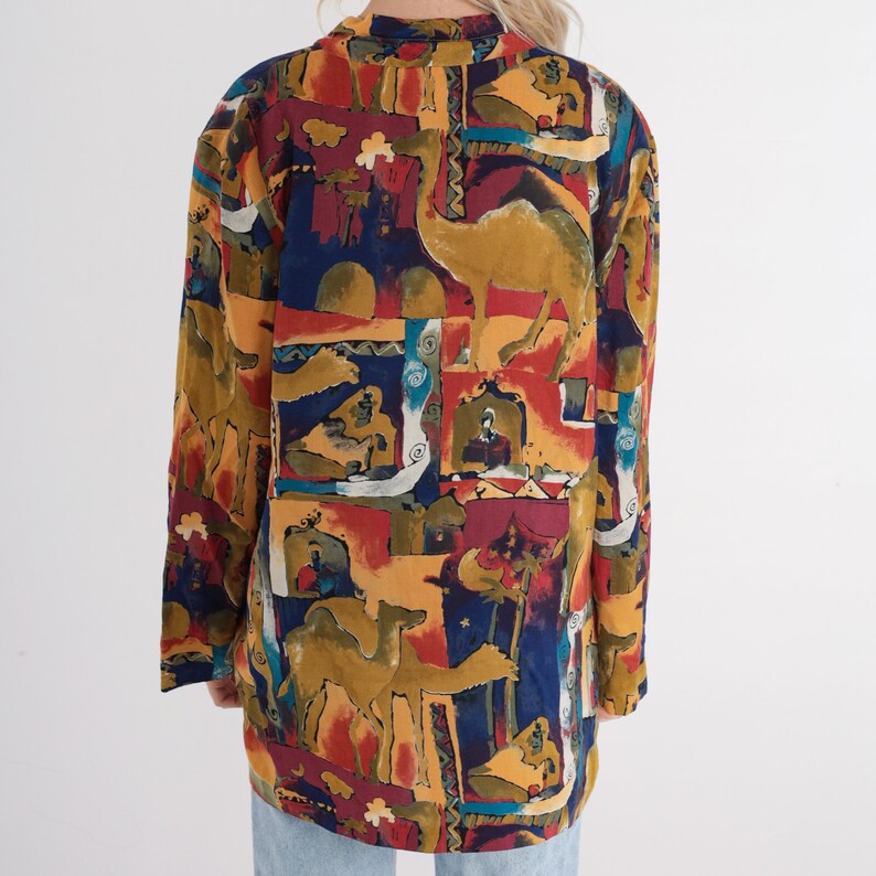 May include: A long-sleeved, oversized blazer with a colorful, patterned design. The pattern features camels, figures, and architectural elements in shades of gold, red, blue, and brown. The blazer has a collared neckline and a loose fit.