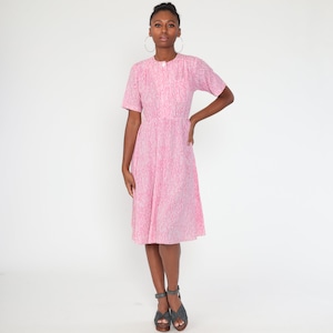 May include: A pink and white patterned dress with short sleeves and a button-up front. The dress has a fitted waist and a flared skirt. The model is wearing gray sandals and large hoop earrings. The background is plain white.