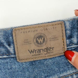 May include: Close-up of blue denim jeans with a brown leather patch. The patch reads "Wrangler AUTHENTIC JEANS" with a circular logo and the words "PREMIUM QUALITY". The jeans are a light blue wash.