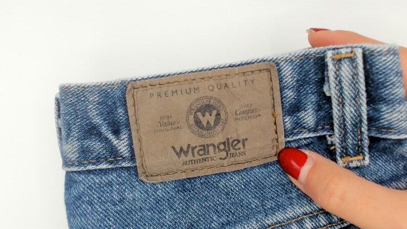 May include: Close-up of blue denim jeans with a brown leather patch. The patch reads "Wrangler AUTHENTIC JEANS" with a circular logo and the words "PREMIUM QUALITY". The jeans are a light blue wash.