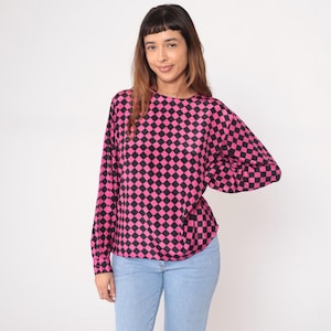 May include: A long-sleeved top featuring a bold checkerboard pattern in black and pink. The top has a round neckline and is paired with light blue jeans. The overall style is casual and eye-catching.