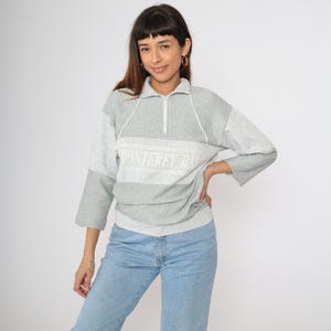 May include: A gray and white striped sweatshirt with a quarter-zip neckline and the text "Monterey Bay" across the chest. The sleeves are a solid gray color. The model is wearing light blue jeans.