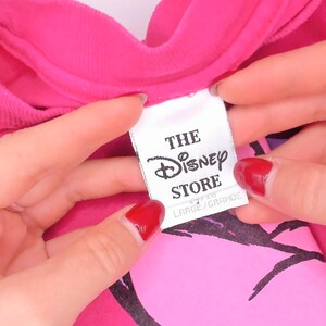 May include: A bright pink t-shirt featuring a white fabric tag with black text that reads THE Disney STORE and LARGE/GRANDE. The garment has a crew neckline and a black graphic print visible beneath the tag on the front of the shirt.