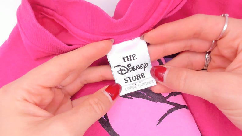 May include: A bright pink t-shirt featuring a white fabric tag with black text that reads THE Disney STORE and LARGE/GRANDE. The garment has a crew neckline and a black graphic print visible beneath the tag on the front of the shirt.