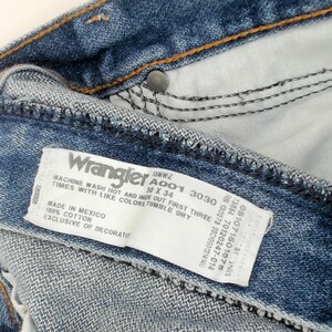 May include: Close-up of blue denim Wrangler jeans. The jeans have a white tag with the Wrangler logo and product information. The tag reads "10 x 34" and "Made in Mexico".