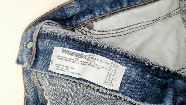 May include: Close-up of blue denim Wrangler jeans. The jeans have a white tag with the Wrangler logo and product information. The tag reads "10 x 34" and "Made in Mexico".