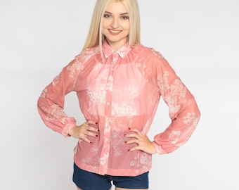 May include: A sheer, long-sleeved, button-down shirt in a soft coral color, adorned with a delicate white floral pattern. The shirt has a classic collar and cuffs, paired with dark blue denim shorts. The model is smiling.